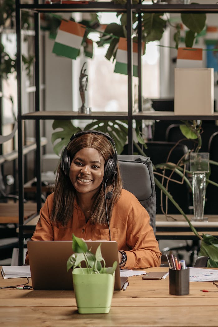 Smiling woman working in call center, wearing headset, surrounded by plants and flags.